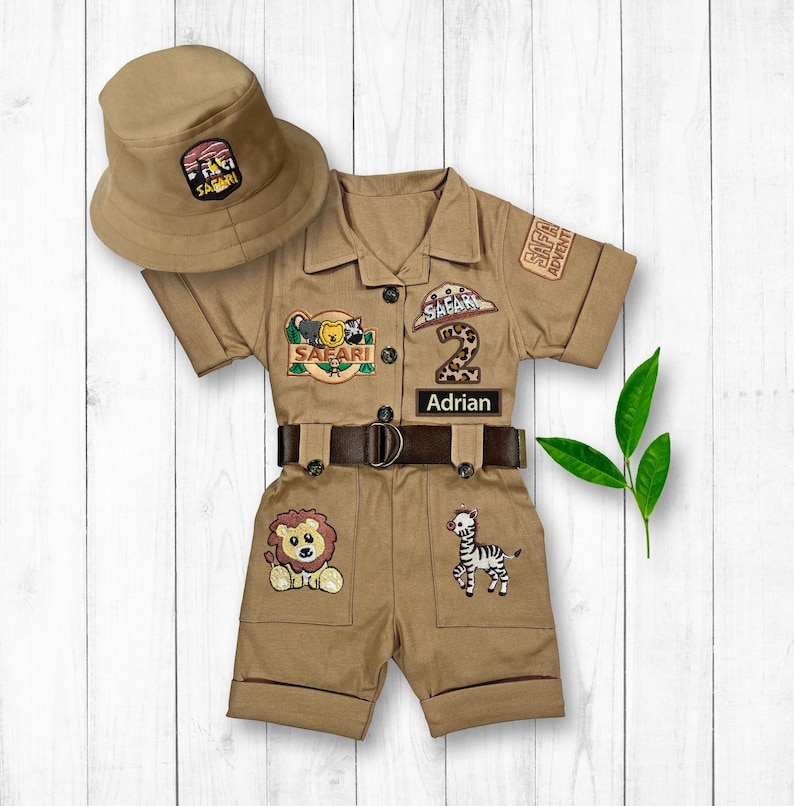 May include: A tan safari-themed outfit for a toddler, featuring a short-sleeved jumpsuit with a brown belt, a safari hat, and various animal and safari-themed patches. The jumpsuit has a patch that says "SAFARI ADVENTURE" and another patch that says "SAFARI". The outfit also has a patch with the number "2" and a name tag that says "Adrian".