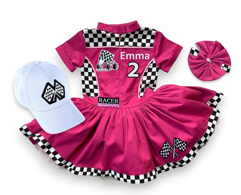 May include: A pink racing-themed outfit set, including a dress, a cap, and a headband. The dress features a checkered pattern, racing flags, and the name "Emma" with the number "2". The white cap also has a checkered flag design.