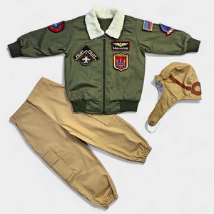 K&ouml;nnte beinhalten: Eine olivgr&uuml;ne Bomberjacke mit wei&szlig;em Kunstfellkragen, verziert mit verschiedenen Aufn&auml;hern zum Thema Luftfahrt sowie den Schriftz&uuml;gen 'Maverick' und 'Pilot'. Das Set enth&auml;lt eine beigefarbene Cargohose und eine passende Fliegerm&uuml;tze.
