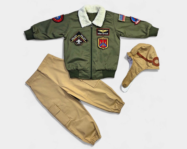 K&ouml;nnte beinhalten: Eine olivgr&uuml;ne Bomberjacke mit wei&szlig;em Kunstfellkragen, verziert mit verschiedenen Aufn&auml;hern zum Thema Luftfahrt sowie den Schriftz&uuml;gen 'Maverick' und 'Pilot'. Das Set enth&auml;lt eine beigefarbene Cargohose und eine passende Fliegerm&uuml;tze.