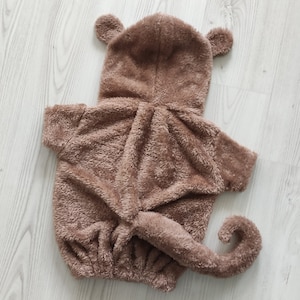 Puede incluir: Un disfraz de mono de peluche marr&oacute;n con capucha y cola. El disfraz est&aacute; hecho de tela suave y peluda.