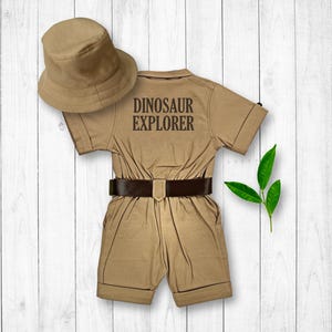 May include: A tan-coloured jumpsuit with short sleeves and a brown belt. The jumpsuit has the text "Dinosaur Explorer" printed on the back. A tan-coloured bucket hat is included.