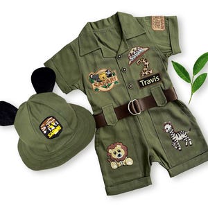 Front view of a personalized olive green safari costume for kids featuring custom "Travis" name and age "2" patches. The set includes a matching green hat with cute black ears, perfect for a Wild One birthday outfit or jungle-themed Halloween costume.
