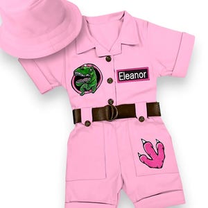 May include: Pink dinosaur costume set with a matching bucket hat. The short-sleeved jumpsuit features a brown belt, a green dinosaur patch, a pink dinosaur footprint, and the name "Eleanor".