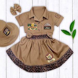 May include: A brown safari-themed dress with a leopard print trim, a matching hat, and a bow. The dress has a patch that says "Safari" and a name tag that says "Lillian".