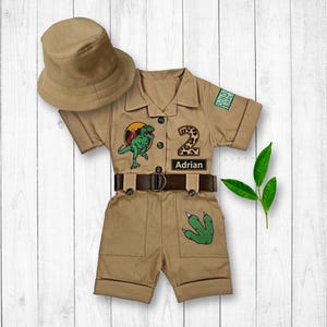 May include: A tan safari-style jumpsuit with short sleeves and a belt. The jumpsuit has patches with a dinosaur, the number 2, and the name "Adrian". It also has a green dinosaur footprint patch. A tan hat is included.