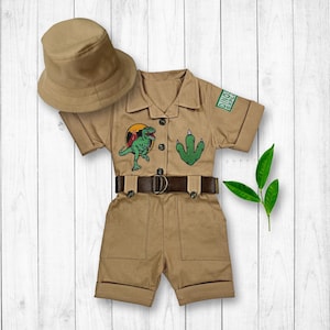 May include: A tan safari-style jumpsuit with short sleeves and a belt. The jumpsuit has a dinosaur and footprint patch on the front and a patch that says "Dino Explorer". The jumpsuit is paired with a tan bucket hat.