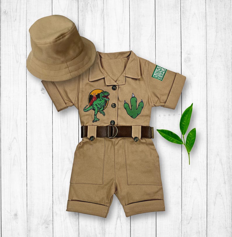 May include: A tan safari-style jumpsuit with short sleeves and a belt. The jumpsuit has a dinosaur and footprint patch on the front and a patch that says "Dino Explorer". The jumpsuit is paired with a tan bucket hat.