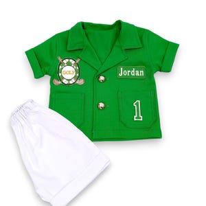May include: A green golf-themed outfit, including a short-sleeved shirt and white shorts. The shirt features a "GOLF" emblem, the name "Jordan", and the number "1". The outfit has silver buttons and two front pockets.