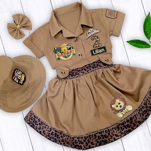May include: A brown safari-themed dress with a leopard print trim. The dress has a matching hat and a patch that says "Safari" and "Lilian".