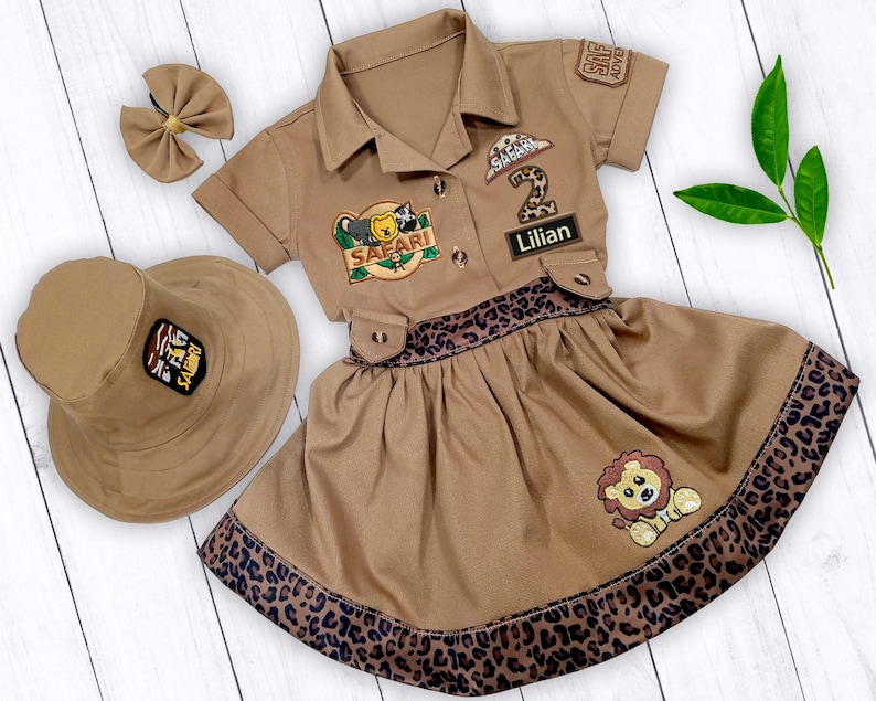 May include: A brown safari-themed dress with a leopard print trim. The dress has a matching hat and a patch that says "Safari" and "Lilian".