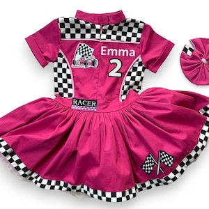 May include: A hot pink racer-themed dress with a matching headband. The dress features a checkered collar and accents, a racing flag graphic, the name "Emma" and the number "2". The skirt has a full, flared design with checkered trim.