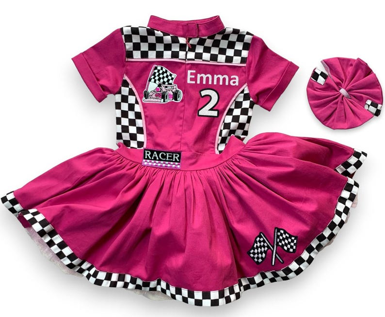 May include: A hot pink racer-themed dress with a matching headband. The dress features a checkered collar and accents, a racing flag graphic, the name "Emma" and the number "2". The skirt has a full, flared design with checkered trim.