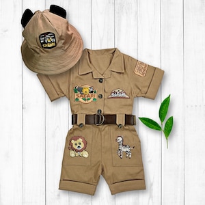 May include: A tan safari-themed jumpsuit with short sleeves and a belt. The jumpsuit has embroidered animal patches, including a lion, zebra, and a bear. The outfit also includes a tan safari hat with black ears.