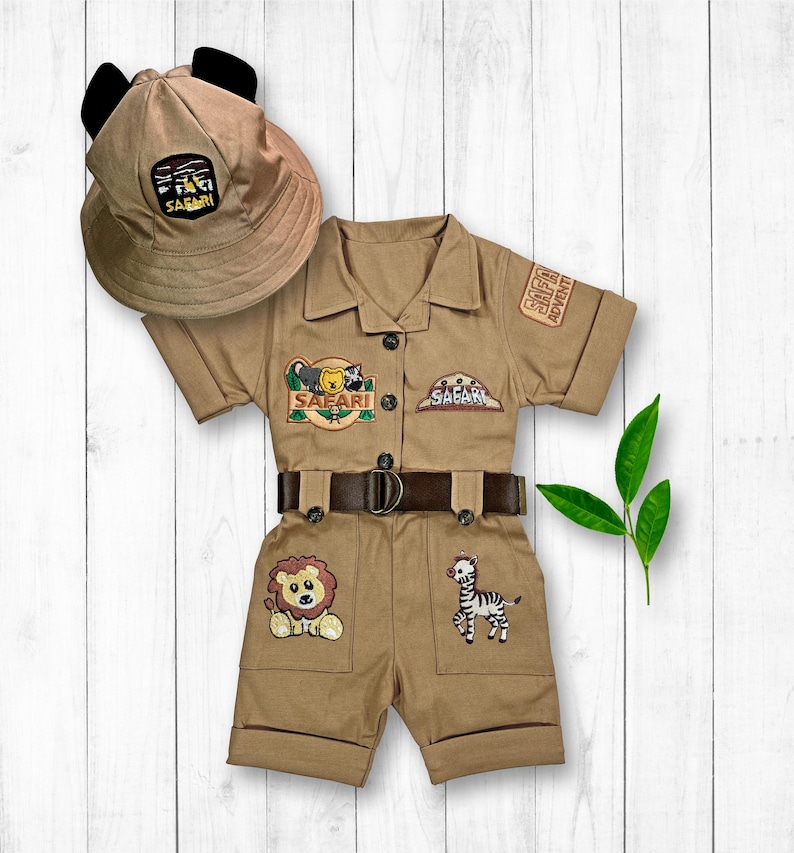 May include: A tan safari-themed jumpsuit with short sleeves and a belt. The jumpsuit has embroidered animal patches, including a lion, zebra, and a bear. The outfit also includes a tan safari hat with black ears.