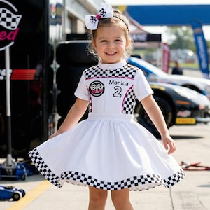May include: A young girl wearing a white racing-themed dress with a checkered pattern on the bodice and hem. The dress features a pink and black racing emblem and the name "Monica" with the number "2". A matching bow adorns her hair.