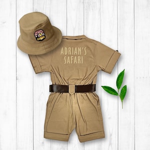 May include: A tan safari jumpsuit with a brown belt and a tan safari hat with a patch that says "Safari". The jumpsuit has the text "Adrian's Safari" embroidered on the back.