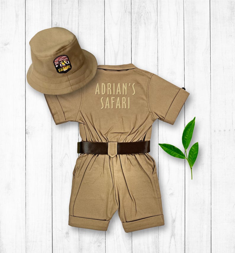 May include: A tan safari jumpsuit with a brown belt and a tan safari hat with a patch that says "Safari". The jumpsuit has the text "Adrian's Safari" embroidered on the back.