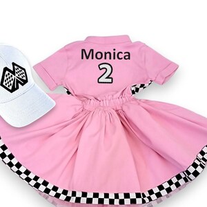 May include: Pink and white checkered dress with a black and white checkered trim. The dress has a white short-sleeved shirt with the name "Monica" and the number "2" on the back. A white baseball cap with a black and white checkered flag design is included.