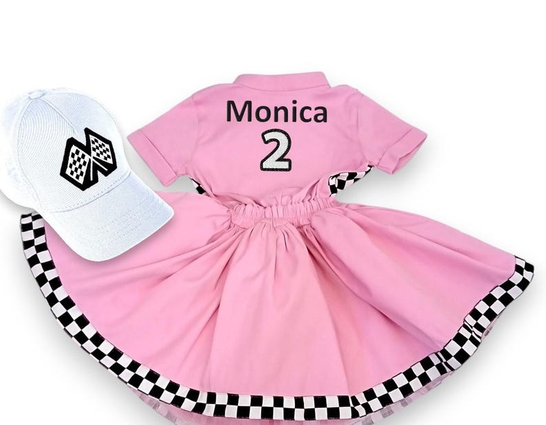 May include: Pink and white checkered dress with a black and white checkered trim. The dress has a white short-sleeved shirt with the name "Monica" and the number "2" on the back. A white baseball cap with a black and white checkered flag design is included.