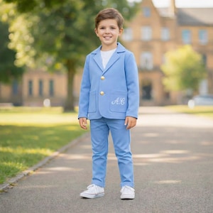 May include: A young person wearing a light blue suit with gold buttons and white sneakers. The suit jacket has pockets and embroidered initials. The outfit is a classic style for special occasions.