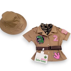 Puede incluir: Un atuendo de safari color canela con un sombrero a juego. El atuendo incluye una camisa de manga corta con parches, un cintur&oacute;n y una etiqueta con el texto "Mina Safari Guide". Se incluye una bufanda con estampado de leopardo.