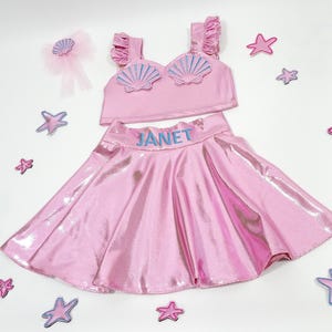 May include: Pink two-piece girls' costume with a seashell top and a sparkly skirt. The top has a blue seashell design and the skirt has the name "JANET" embroidered on the waistband.