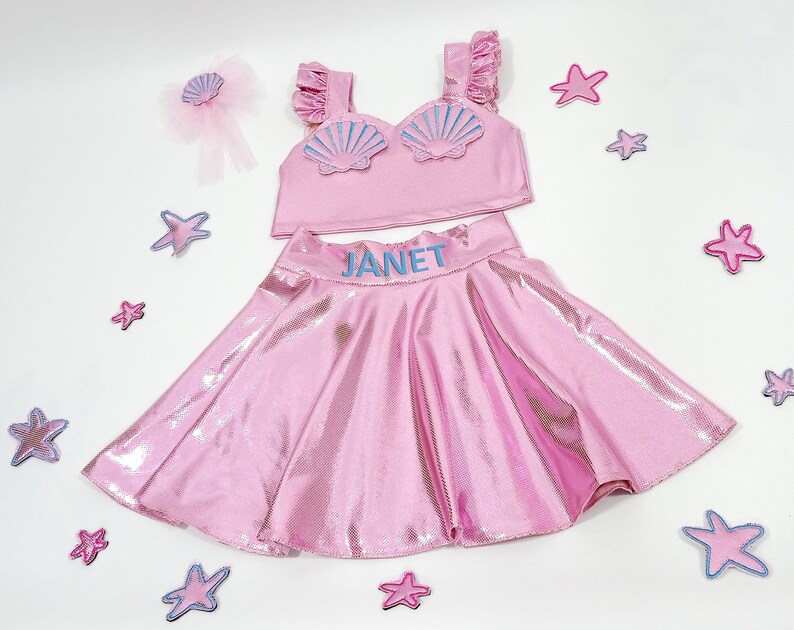May include: Pink two-piece girls' costume with a seashell top and a sparkly skirt. The top has a blue seashell design and the skirt has the name "JANET" embroidered on the waistband.