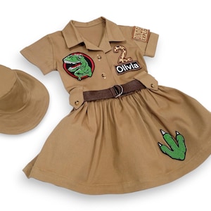 Peut inclure: Ensemble de costume d'explorateur de dinosaures beige avec un chapeau assorti. La tenue comprend une robe chemise à col avec une ceinture marron, des écussons de dinosaures et le nom "Olivia". Une empreinte de pas de dinosaure verte est sur la jupe.