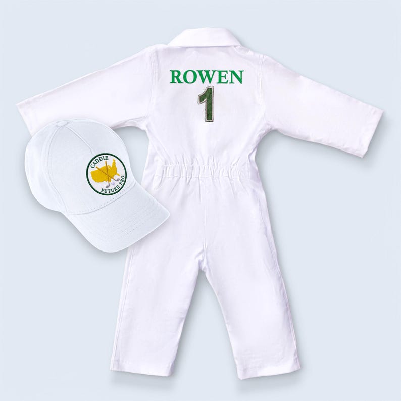 Back view of the personalized kids' jumpsuit highlighting the premium green embroidery. The name "ROWEN" is stitched in large block letters across the shoulders with a large number 1 centered below. This view emphasizes the high-quality customization and professional finish of the embroidery, paired with a matching white cap.