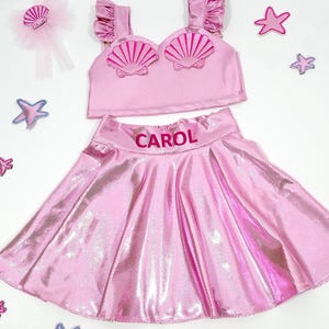 May include: Pink mermaid costume for a child, featuring a sparkly pink skirt with the name "CAROL" embroidered on the waistband, a pink crop top with a shell design, and a pink hair bow with a shell accent.