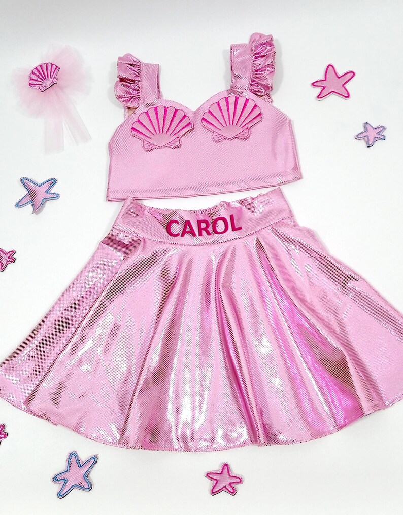May include: Pink mermaid costume for a child, featuring a sparkly pink skirt with the name "CAROL" embroidered on the waistband, a pink crop top with a shell design, and a pink hair bow with a shell accent.