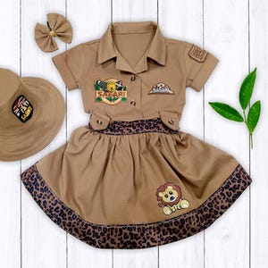 May include: A brown safari-themed dress with a lion and a safari hat patch. The dress has a brown and black leopard print trim and a matching brown hat.