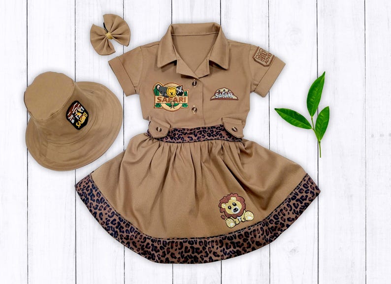 May include: A brown safari-themed dress with a lion and a safari hat patch. The dress has a brown and black leopard print trim and a matching brown hat.