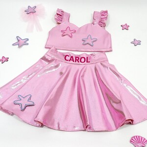 May include: Pink two-piece outfit with a sparkly skirt and a crop top featuring a star design. The skirt has the name "CAROL" embroidered on the waistband.