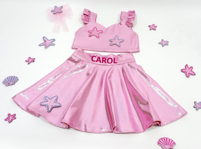 May include: Pink two-piece outfit with a sparkly skirt and a crop top featuring a star design. The skirt has the name "CAROL" embroidered on the waistband.