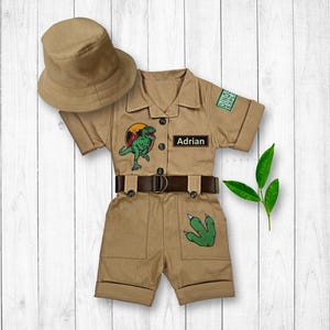 May include: A tan safari jumpsuit with a dinosaur patch, a brown belt, and a green dinosaur footprint patch. The jumpsuit has a name tag that says "Adrian" and a patch that says "Dino Explorer". A tan safari hat is included.