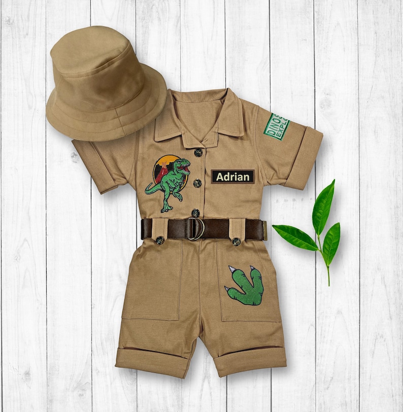 May include: A tan safari jumpsuit with a dinosaur patch, a brown belt, and a green dinosaur footprint patch. The jumpsuit has a name tag that says "Adrian" and a patch that says "Dino Explorer". A tan safari hat is included.