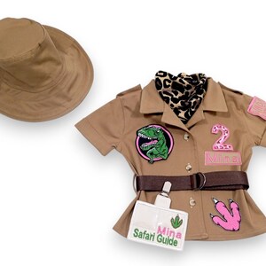 Puede incluir: Un conjunto de safari color canela con un sombrero a juego. La camisa tiene un parche de dinosaurio, un parche rosa con el n&uacute;mero 2 y una etiqueta con el nombre "Mina Safari Guide". Un cintur&oacute;n marr&oacute;n y una bufanda con estampado de leopardo completan el look.