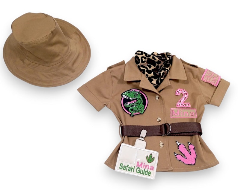 Puede incluir: Un conjunto de safari color canela con un sombrero a juego. La camisa tiene un parche de dinosaurio, un parche rosa con el n&uacute;mero 2 y una etiqueta con el nombre "Mina Safari Guide". Un cintur&oacute;n marr&oacute;n y una bufanda con estampado de leopardo completan el look.
