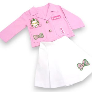May include: A pink blazer with a golf-themed patch, a bow, and a name tag that says "Emma". The blazer is paired with a white skirt with a bow patch.