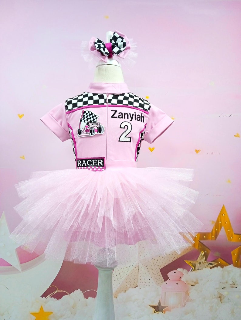 May include: Pink and black checkered racing-themed tutu outfit for a child. The outfit includes a pink and black checkered top with a pink race car graphic, the name "Zanyiah" and the number "2" printed on the front. The top is paired with a pink tulle tutu.