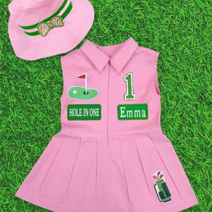 May include: Pink golf-themed outfit set, including a sleeveless dress and a sun hat. The dress features a pleated skirt, a collared neckline, and patches with the text "HOLE IN ONE" and "Emma". The hat has green and white striped accents.