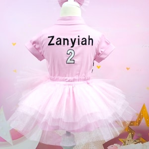 May include: Pink tutu dress with a pink short-sleeved shirt that says "Zanyiah 2" on the back. The dress is on a white mannequin with a black bow on top of its head.