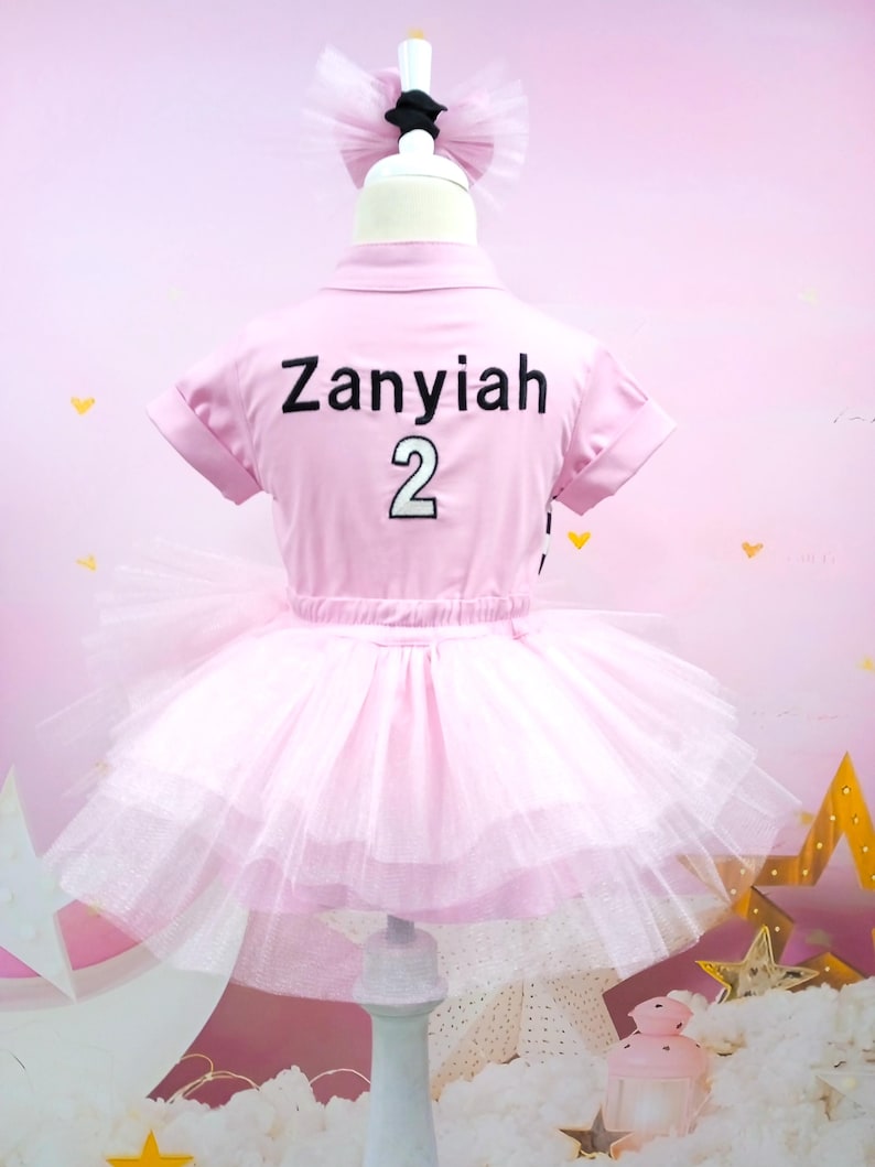 May include: Pink tutu dress with a pink short-sleeved shirt that says "Zanyiah 2" on the back. The dress is on a white mannequin with a black bow on top of its head.