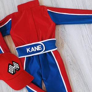 May include: A red and blue racing suit with white accents and the name "KANE" on the belt. A matching red baseball cap with a checkered flag design is also included. The suit has a high collar and long sleeves.