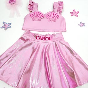 May include: Pink mermaid costume for a child with a shell top and a skirt. The skirt has the name "CAROL" embroidered on it.
