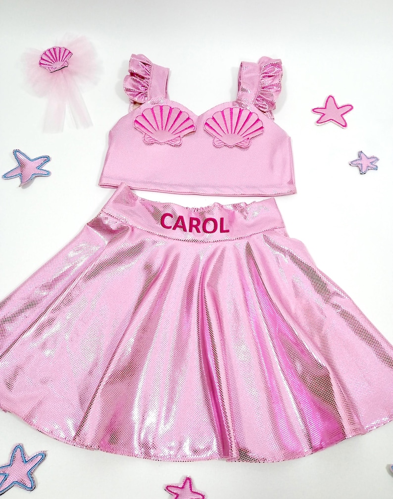 May include: Pink mermaid costume for a child with a shell top and a skirt. The skirt has the name "CAROL" embroidered on it.