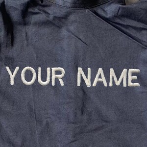 May include: A dark blue jacket with white piping on the sleeves and a white embroidered "YOUR NAME" on the back.
