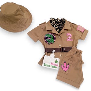 Puede incluir: Un conjunto de disfraz de safari color canela para ni&ntilde;os, que incluye una camisa con botones, pantalones cortos y un sombrero de ala ancha. La camisa presenta un parche de dinosaurio, un parche con el n&uacute;mero 2 y un cintur&oacute;n marr&oacute;n. Los pantalones cortos tienen un parche rosa con una huella de dinosaurio.